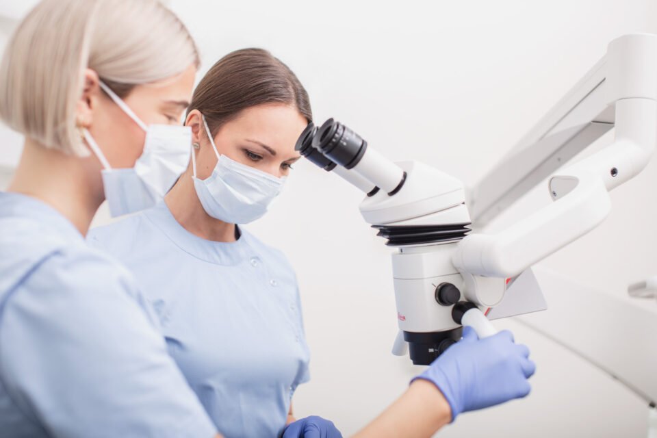 „Evadenta“ klinikos specialistės dirba su odontologiniu mikroskopu.